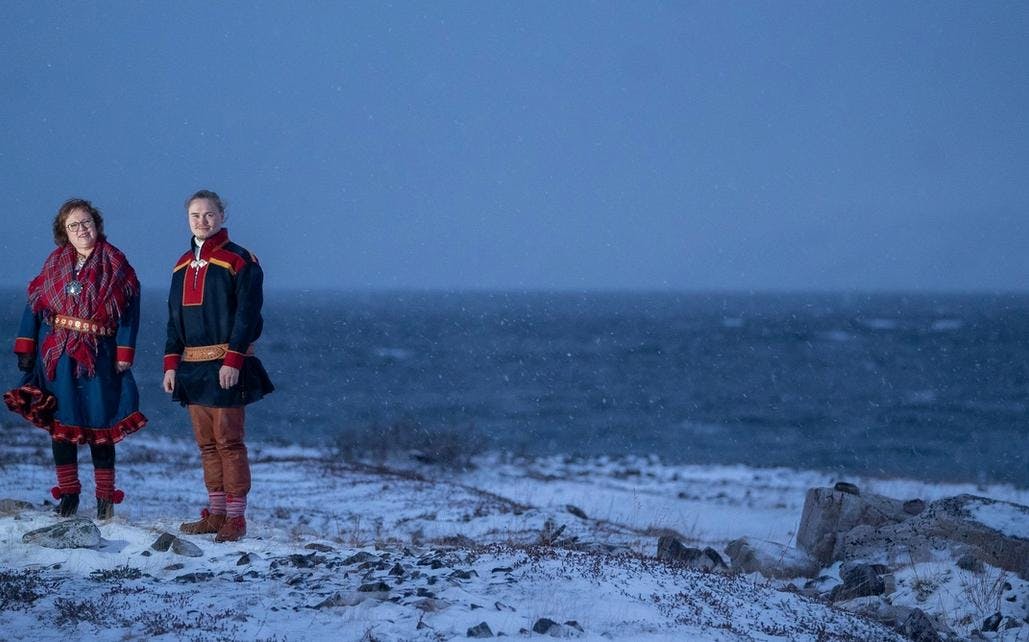 Et bilde av to artister i samiske kofter som står i et åpent landskap foran havet.